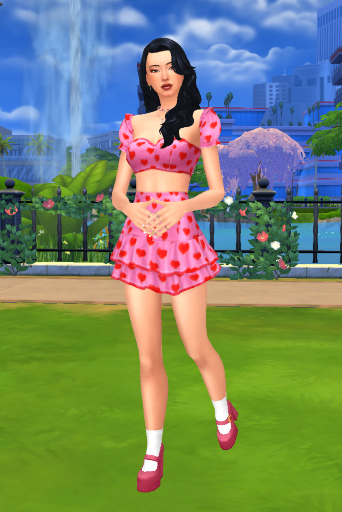 A female Sim wearing a Sims 4 CC outfit stands in a park. She is dressed in a matching pink two piece set with red hearts all across the fabric. She has on a pair of pink platform heels and white socks.