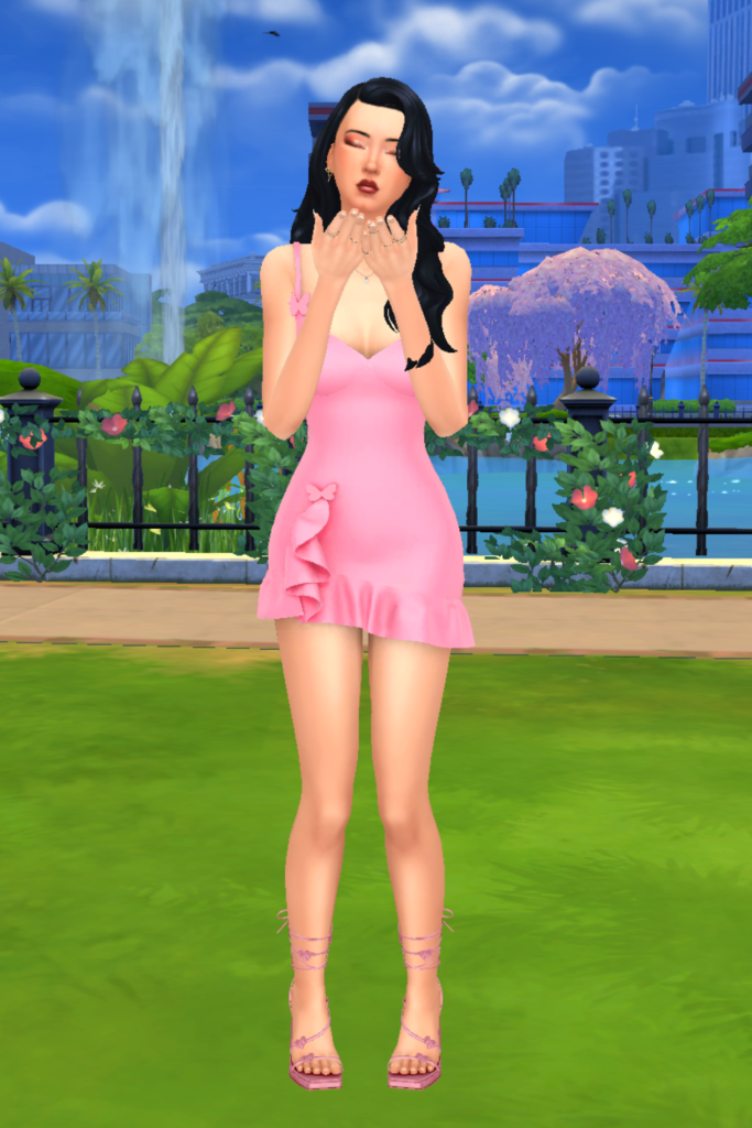 A female Sim wearing a Sims 4 CC outfit stands in a park. She is dressed in a pink mini dress. The dress has ruffles along the hem and some pink butterflies throughout. She has on a pair of pink strappy heels that have hearts along the straps