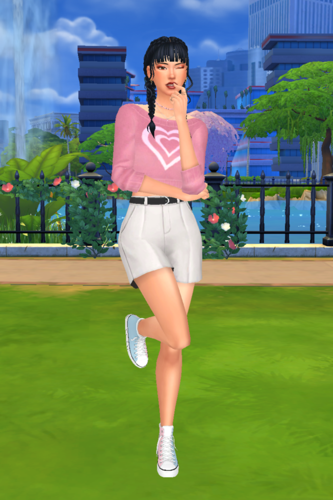 A female Sim wearing a Sims 4 CC outfit stands in a park. She is dressed in a cute pink cropped sweater with a white heart in the centre. She also has on a pair of white high-waisted dress shorts and a pair of pink, blue, and white coloured converse.